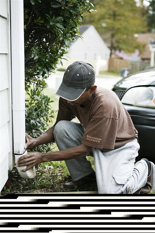 Man kneeling near an exterior wall adjusting a plastic downspout extension on a house (FEMA photo by Andrea Booher)